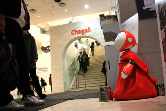 Imagen de DONA en perspectiva, un robot con una capa roja y cara blanca con una lata de donaciones frente a él.